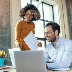 A man, sitting and a woman, standing look over documents and the screen of a computer whilst smiling