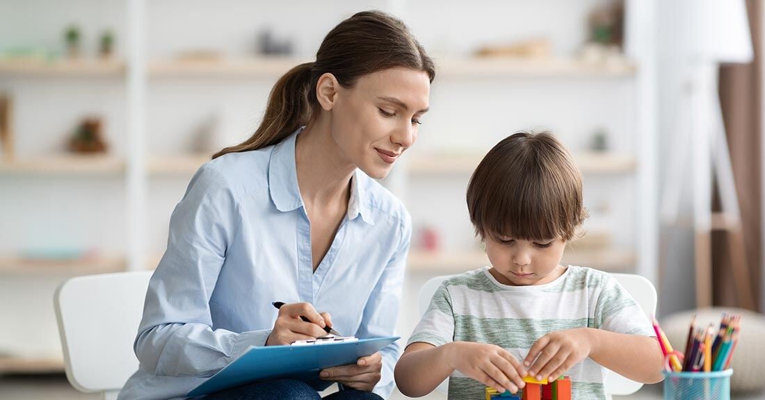 Female mental health worker with a notepad and pen, overseeing a five-year old child performing a task with blocks.