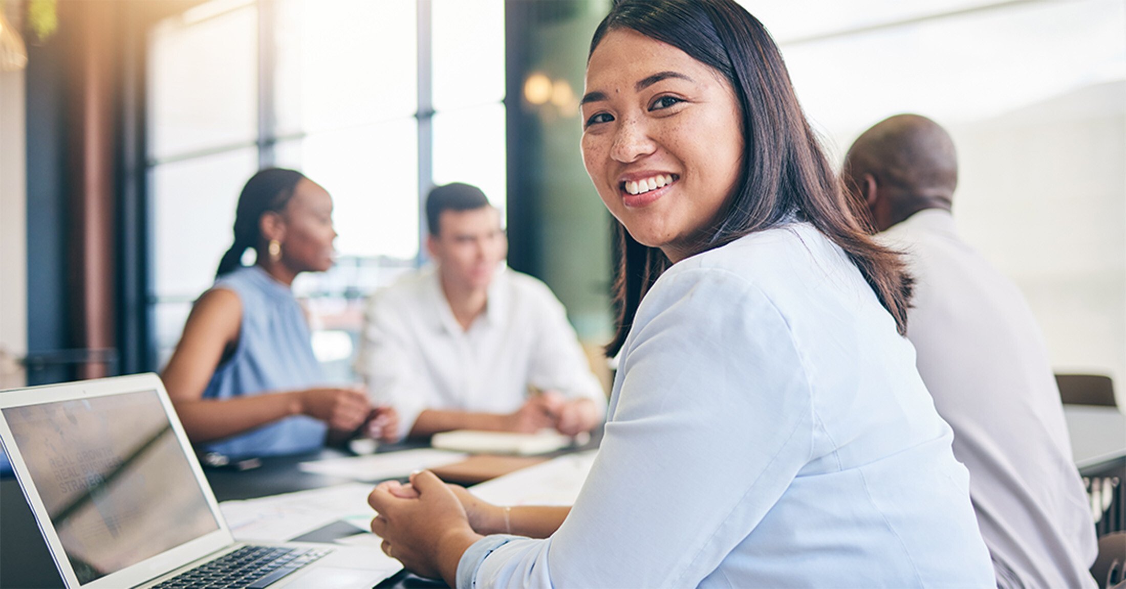 A woman seated at a conference table with her coworkers smiling at the camera while using a laptop.